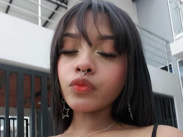 HannaCrow, com cabelo preto, olhos fechados, piercing no nariz, brincos de estrela e batom vermelho, fazendo um biquinho, vestindo uma blusa preta com alças, parecendo fofa e sexy..