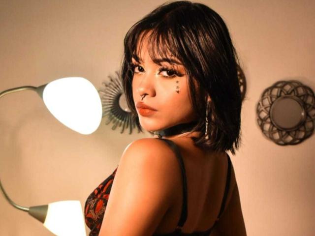 HannaCrow with short black hair, brown eyes, nose piercing, and septum ring, wearing a black and red lace bra, looking over shoulder, standing in front of a lamp.