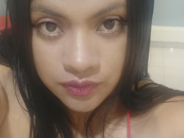 DahianaColeman's face, brown eyes, black straight hair, pink lipstick, slight pout, red spaghetti strap visible, soft light, slight blush on cheeks, centered in the image.