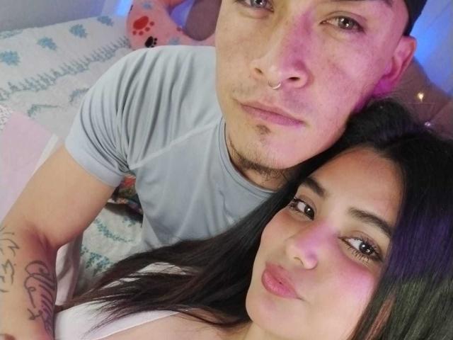TommyAndGrace with a nose ring, grey t-shirt, and black hair, next to TommyAndGrace with long black hair, pink lips, and a white top, in a bedroom.