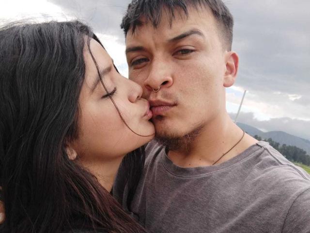 TommyAndGrace kissing, Tommy's nose pierced, short brown hair, stubble, gray shirt; Grace's long black hair, closed eyes, Tommy's slightly open eyes, cloudy sky in background.