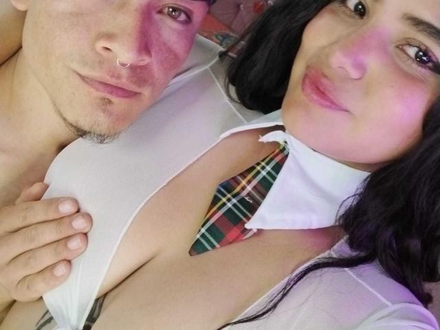 TommyAndGrace with pink cheeks, nose ring, and slight stubble, holding white shirt to expose TommyAndGrace's large tits, wearing a colorful plaid tie, and smiling.