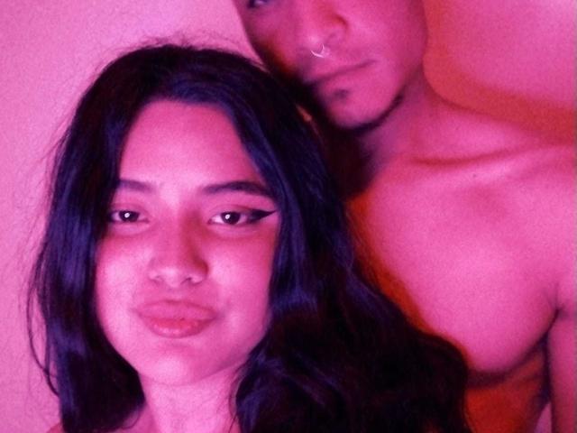 TommyAndGrace with dark hair, nose ring, and eyeliner, standing behind TommyAndGrace, shirtless, with a nose ring and a slight smile, in pink lighting.