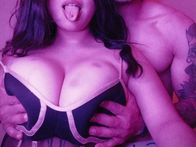 TommyAndGrace's big tits, in a black bra with pink trim, are being held by Tommy's tattooed hands. She's sticking her tongue out. Purple lighting.