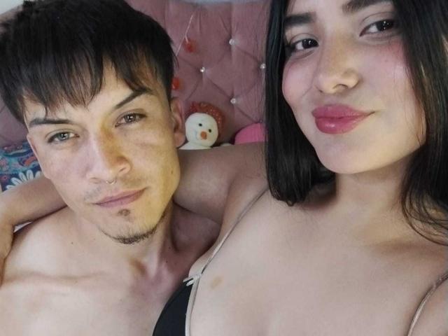 TommyAndGrace with short brown hair and a nose ring, shirtless, next to TommyAndGrace with black hair and pink lipstick, showing small tits, in a bed.