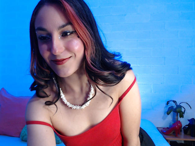 AmyStreetwise69 with red and black wavy hair, white shell necklace, red off-shoulder top, smiling, light skin, subtle makeup, red lipstick, sitting, blue background, small potted plant on right.