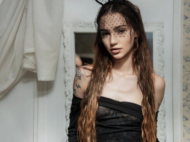 SophieKitty con cabello castaño ondulado, vestido negro con un hombro descubierto, tocado de malla con lunares, tatuaje en el hombro derecho, pared blanca y cortina blanca en el fondo. Sus pechos son pequeños..