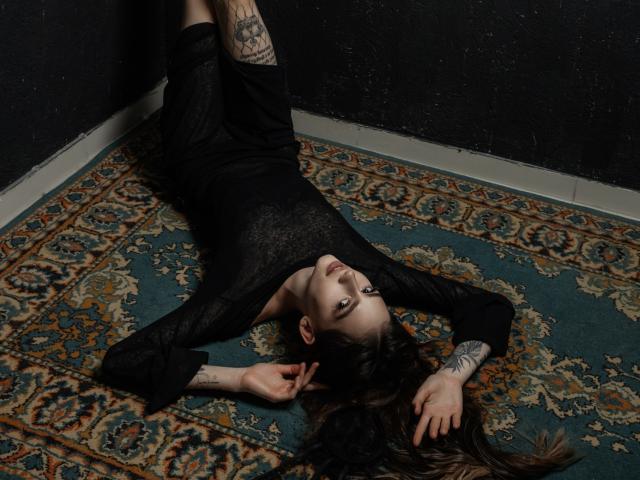 SophieKitty está deitada de costas, com os braços acima da cabeça, vestindo um vestido preto transparente de manga comprida, que revela tatuagens em seu braço direito e perna, sobre um tapete ornamentado..