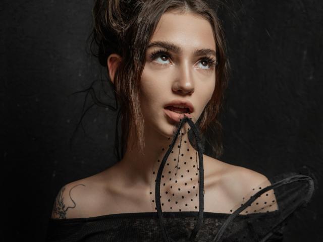 SophieKitty, con i capelli scuri, che tiene in bocca un tessuto nero a pois, indossa una maglietta nera con una spalla scoperta, ha un tatuaggio sulla spalla sinistra, occhi marroni intensi, sfondo scuro..