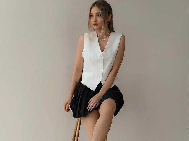 EvaCaroline ha i seni coperti da un gilet bianco senza maniche, una gonna nera, e una gamba appoggiata su uno sgabello di legno, indossa una collana dorata, ha i capelli castano chiaro e un'espressione neutra..