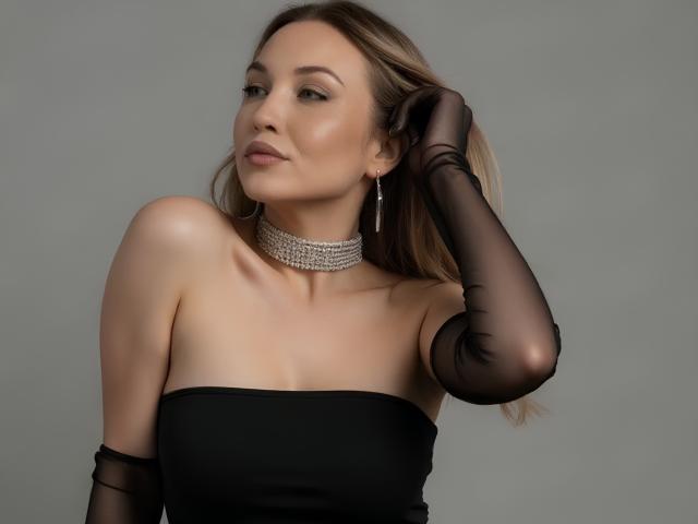 EvaCaroline con i capelli biondi, un abito nero senza spalline, un guanto nero trasparente, una collana di diamanti e orecchini, che appare compiaciuta con una mano tra i capelli, mostrando il seno..