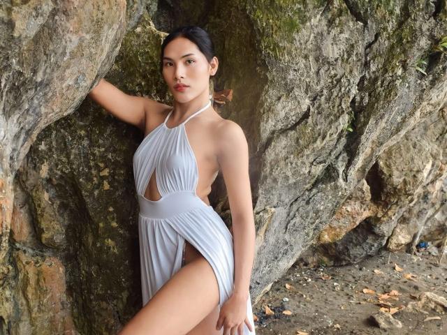 SofiaCumness wearing a white halter dress with a cutout, standing against a rocky wall, showing her ass and a side of her pussy. She has dark hair, tan skin.