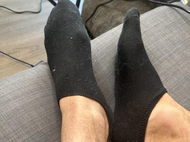 OnlyRoro's legs in black socks, slightly dirty, with visible hairy calves, resting on a gray couch. Socks cover toes, showing slight wear. Background includes wooden floor.