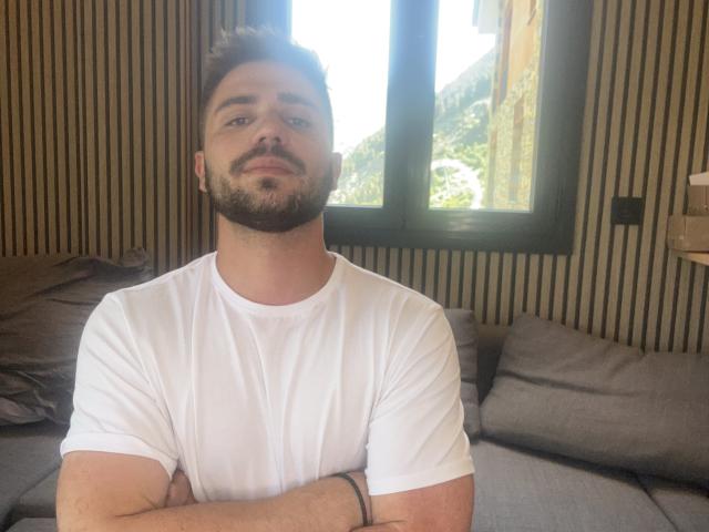 OnlyRoro with a trimmed beard and short dark hair, wearing a white t-shirt, arms crossed, looking at the camera, standing in front of a window with green outside.