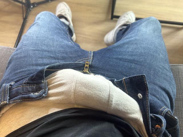 Le jean bleu de OnlyRoro est ouvert, révélant une chemise blanche et un sous-vêtement beige, avec des baskets grises sur un plancher en bois. La perspective est au niveau de son entrejambe..
