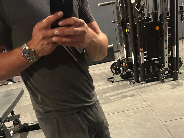 Les bras musclés de OnlyRoro, portant un t-shirt gris et un short assorti, prennent un selfie avec un téléphone dans une salle de sport. Il porte une montre argentée brillante à son poignet gauche..