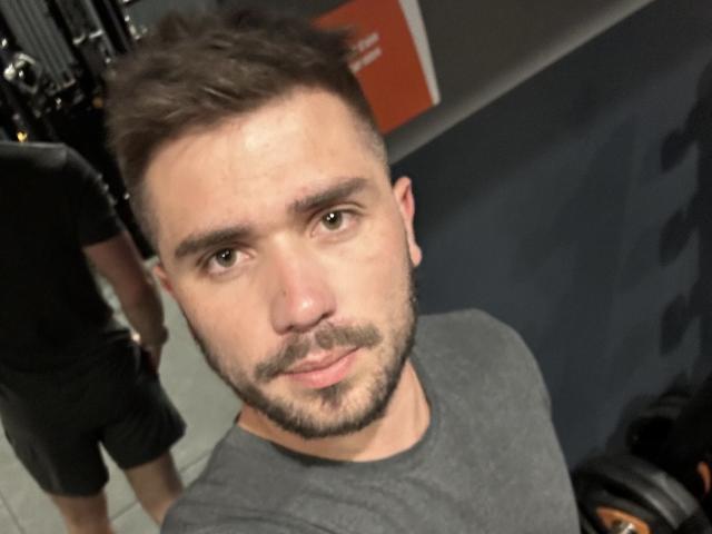 OnlyRoro, avec une courte barbe et moustache brunes, un t-shirt gris, regardant la caméra, debout dans une salle de sport, avec un short noir d'une autre personne visible en arrière-plan..