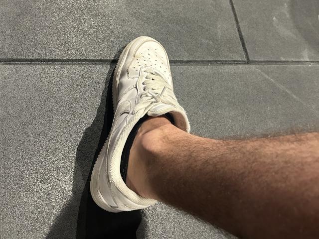 Het harige been van OnlyRoro, in een witte sneaker met zwarte zolen, staat op een grijze tegel. De sneaker is veters, de voet is lichtjes gekanteld, er is een schaduw op de tegel..