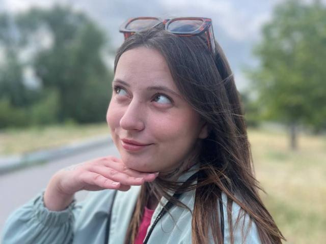 LilaVame has brown hair, glasses on her head, a light green jacket, and a pink shirt. She's smiling slightly, looking to the side with her hand under her chin.