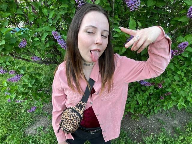 LilaVame con una camisa rosa, un bolso de leopardo, la lengua afuera, señalando hacia arriba, hojas verdes, flores moradas, cabello castaño, piel blanca, collar, anillo, hierba, al aire libre, ligeramente despeinada..