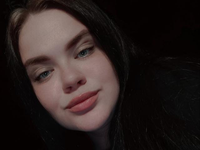 AnnLaura has pale skin, blue eyes, and full pink lips. She has dark brown, slightly wavy hair, and a subtle, confident smile. Her eyebrows are neatly shaped.