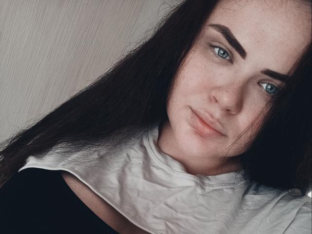 AnnLaura has pale skin, blue eyes, dark brown eyebrows, long black hair, and a slight smile. She's wearing a black top with a white collar.