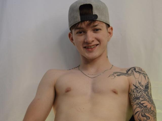 JakeDavins, sin camisa y con un gorro gris, sonríe. Tiene un brazo derecho tatuado, pezones pequeños y un collar de cadena. Su piel es clara y tiene perforaciones en las orejas..