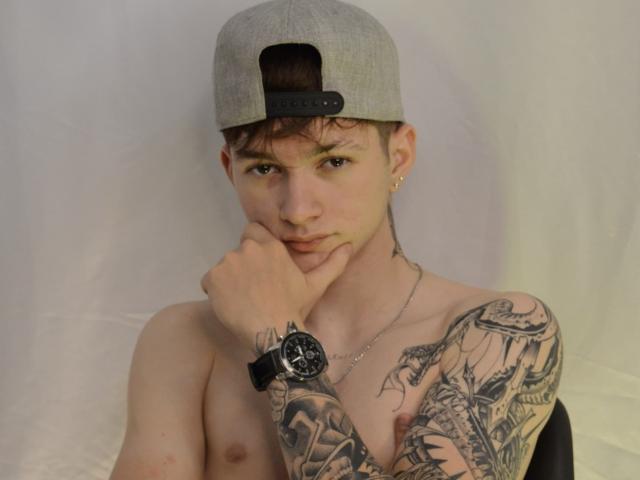 JakeDavins, sin camisa, con un gorro gris, un reloj negro y aros dorados, con tatuajes en su brazo derecho, y la mano cerca de la boca, mirando a la cámara..