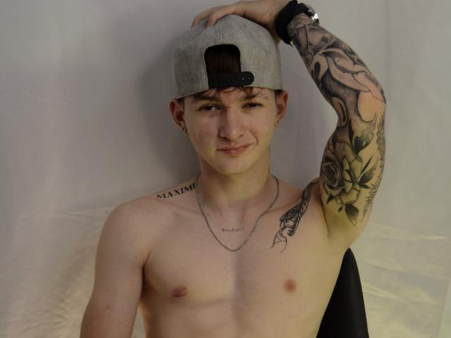 JakeDavins ohne Hemd, mit einer grauen Baseballkappe, tätowierter rechter Arm, trägt eine Halskette, lächelt, Hand auf dem Kopf, Armband am Handgelenk, helle Haut, kleine Brustwarzen, sitzt auf einem schwarzen Stuhl..