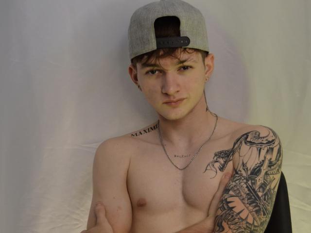 JakeDavins zonder shirt, met een grijze achterstevoren pet, kleine tepels en een gedetailleerde tatoeage op zijn rechterarm, draagt een ketting, heeft zijn armen gekruist en heeft een lichte grijns..