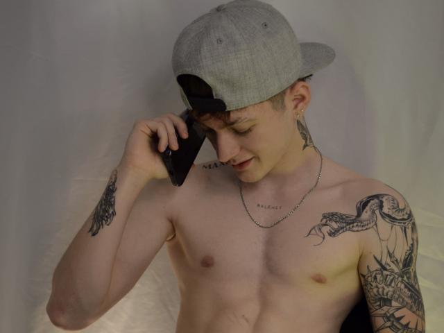 Shirtless JakeDavins with tattoos on arms and chest, wearing a gray baseball cap, holding a black phone to his ear, looking down, silver chain necklace.