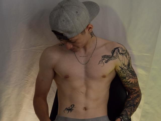 JakeDavins sin camisa, con tatuajes, usando una gorra gris, mirando hacia abajo, mostrando un pecho blanco, delgado y tonificado, con tatuajes y un tatuaje de serpiente en su brazo derecho..