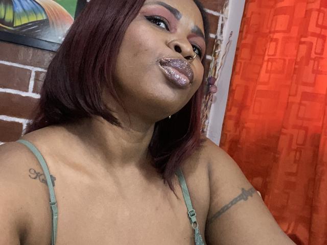AshleyPumper, les lèvres brillantes d'un rouge violet foncé, les cheveux d'un rouge profond, un soutien-gorge vert, des tatouages sur les bras, un anneau au nez, et une légère moue, devant un rideau rouge et un mur de briques..