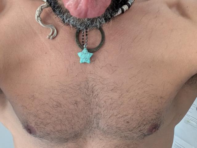 SophiaStorm, avec ses cheveux foncés, sa petite poitrine et son corps mince, portant des bas noirs en résille et un string noir, est allongée sur un lit gris et tient une télécommande..