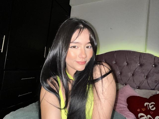ValerieSweet69 in posizione accovacciata, indossa una maglia nera trasparente che rivela una coscia tatuata con un disegno dettagliato che raffigura un volto, lunghi capelli scuri, eyeliner nero, guarda verso la fotocamera, fotografia in scala di grigi..