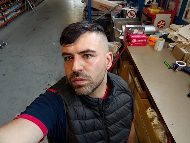 Vio69x a un visage sérieux, des cheveux courts et foncés, et une barbe, et porte un gilet noir matelassé par-dessus une chemise rouge et noire, dans un atelier encombré..