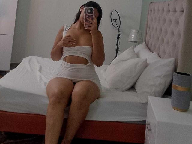 AshleyRosel está sentada en una cama, vestida con un vestido blanco con aberturas, cubriéndose los senos con una mano, mostrando su trasero, y se está tomando una selfie con su teléfono. El escenario es un dormitorio..