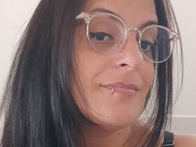 Julires, com cabelo preto e liso, usa óculos redondos, tem um piercing no lábio e um leve sorriso. Seu nariz é achatado e tem uma pequena pinta abaixo dele..