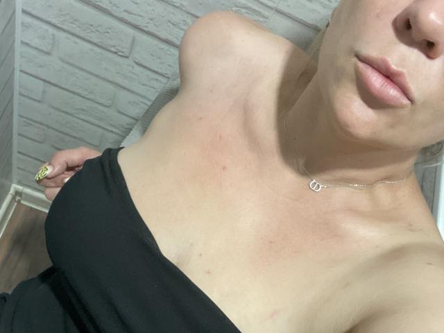 Het bovenlichaam van AnnaMilc, waarbij haar blote schouders en zwarte bandjesjurk zichtbaar zijn, met haar hand op haar zij en gele nagels, draagt ze een kleine ketting. Haar borsten zijn lichtelijk zichtbaar..