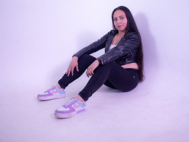 AliceBlackx, zittend met gebogen benen, draagt een zwarte leren jas, een wit T-shirt, zwarte jeans, paars-witte sneakers, lang zwart haar, een lichte glimlach, handen op de knieën, witte achtergrond..