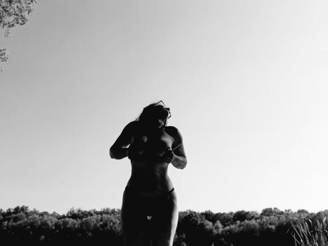 Schwarz-weißes Foto im Silhouettenstil von RivieraM, die rennt, mit geneigtem Kopf, Hände in der Nähe der Brust, in einem Bikini-Unterteil, vor einem hellen Himmel mit einer Baumlinie im Hintergrund..