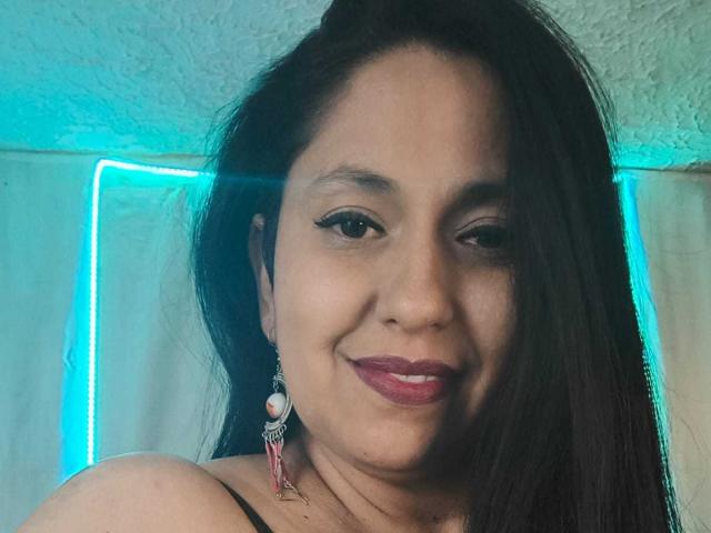 HolyHottie ha una pelle marrone media, capelli neri ed è sorridente con un rossetto rosa. Indossa una canotta nera con spalline e orecchini decorativi con elementi rosa e bianchi..