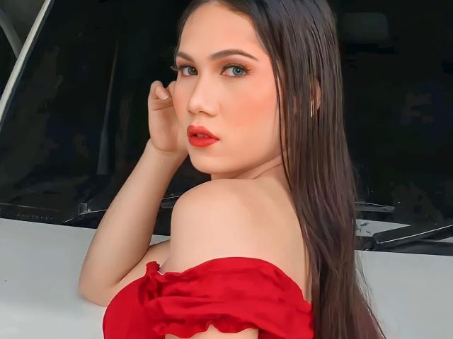Amiraheather, com longos cabelos castanhos, batom vermelho e uma blusa vermelha com um ombro descoberto, olhando por cima do ombro, expondo seu ombro nu e parte superior do peito. Ela está em um carro branco..
