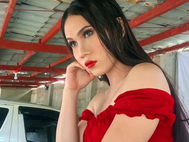 Amiraheather, avec de longs cheveux noirs, une robe rouge à une épaule, du rouge à lèvres rouge et un maquillage foncé, touchant sa joue dans un garage avec des poutres rouges au plafond et une camionnette blanche..