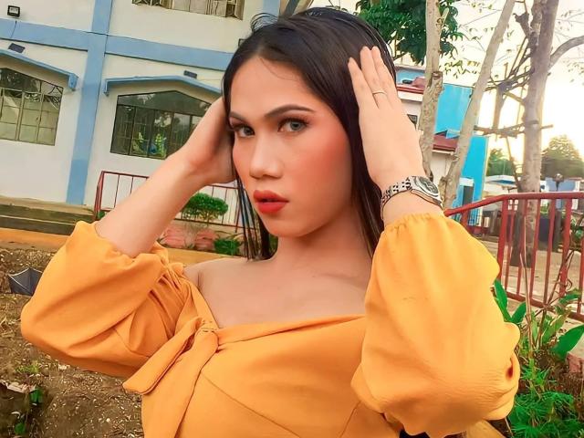 Amiraheather with hands in her black hair, wearing an off-shoulder orange dress, looking at the camera with red lips, small nose, and brown eyes, showing her tits slightly.