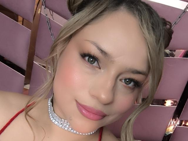 AgathaWalker with blonde hair, blue eyes, pink lipstick, and a diamond choker, smiling at the camera, showing off her boobs in a red top.