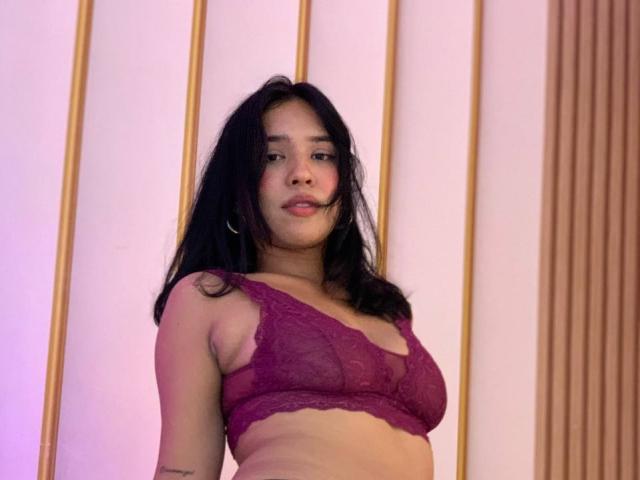 Cinnamon : Filles à la peau marron moyen, aux cheveux noirs, portant un soutien-gorge en dentelle violette transparent, montrant ses petites seins et un tatouage discret sur son bras gauche, regardant légèrement sur le côté..