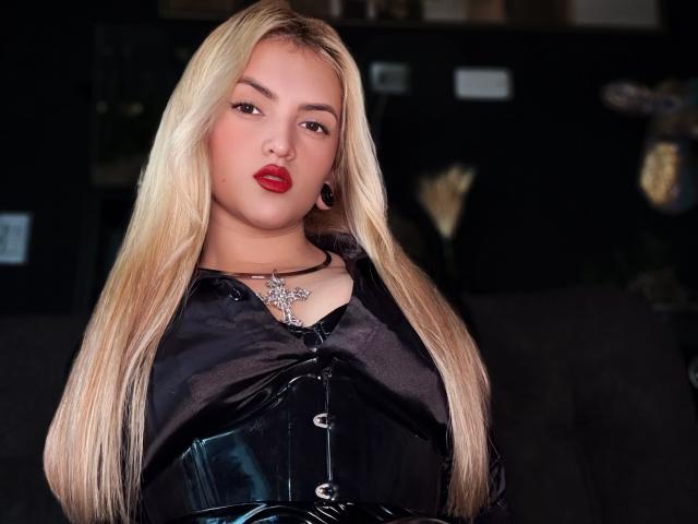 Blond FiorellaNox with red lips, black shiny leather corset, and cross necklace, has long hair, black ear rings, and a serious expression, standing in dimly lit room.