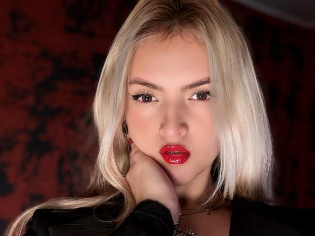 FiorellaNox, rubia, con labios rojos brillantes, delineador de ojos oscuro y piel clara, sosteniendo su cuello con manos vestidas de negro, mirando fijamente a la cámara..