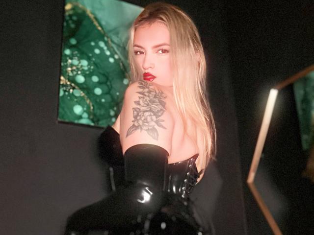 FiorellaNox, rubia, con lápiz labial rojo, guantes de látex negros y un tatuaje floral en el hombro, mirando por encima del hombro, mostrando sus pechos, en una habitación oscura..